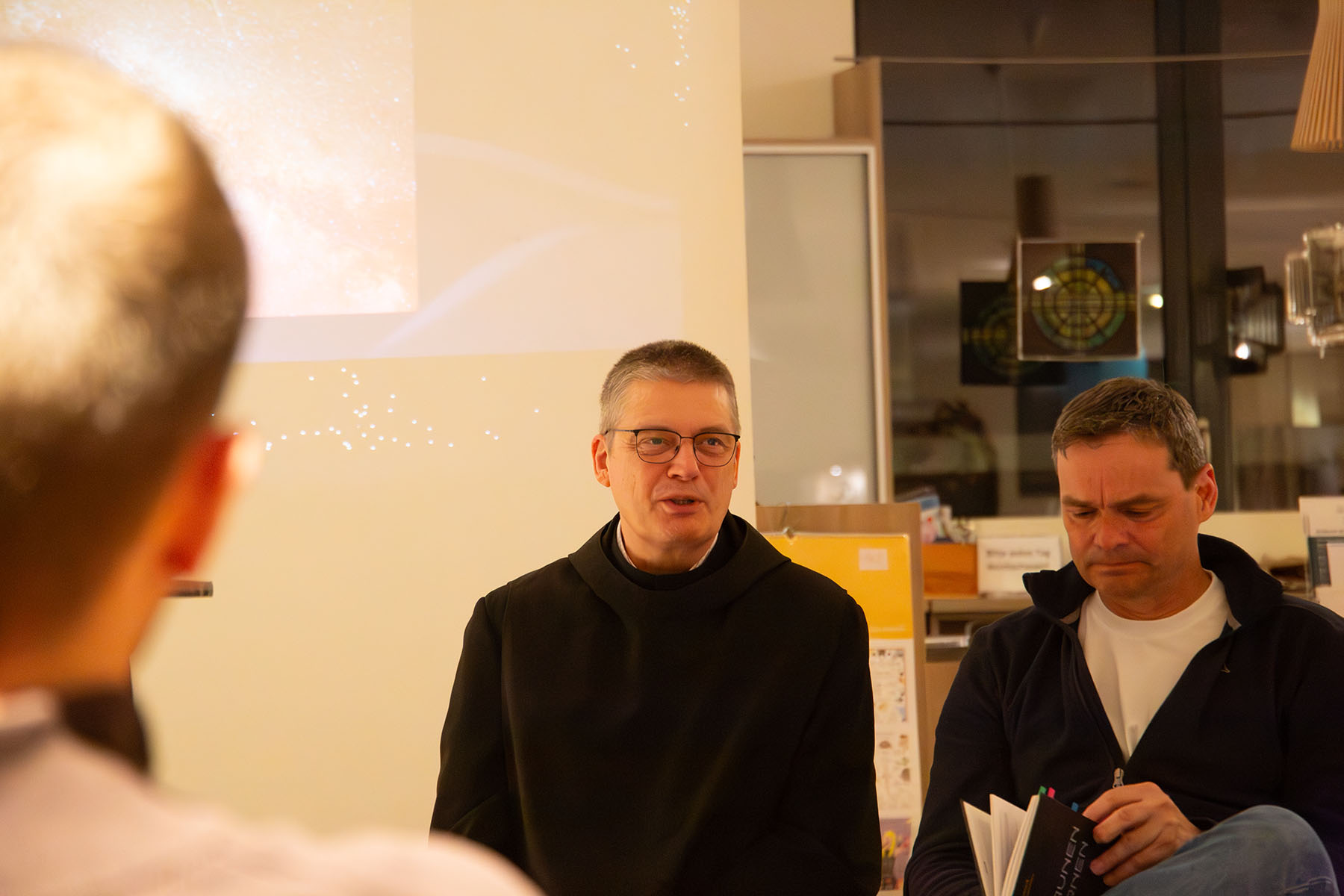 Mann im Habit erzählt, Mann daneben schaut in ein Buch